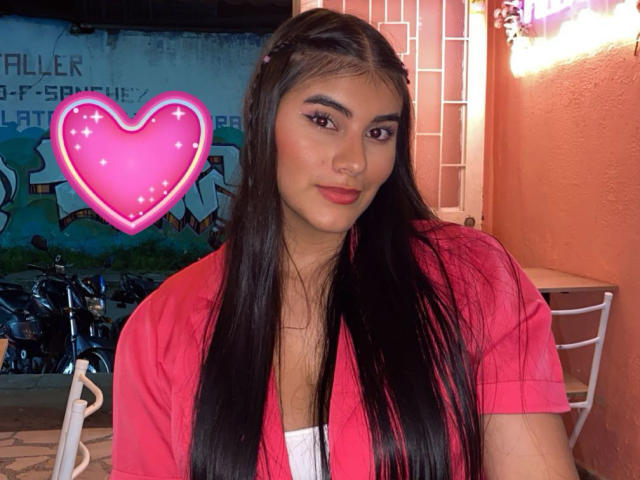 AbbyKell heeft lang zwart haar, een rood shirt, en een roze hart-emoji boven haar borsten. Ze zit met een zelfverzekerde glimlach, met make-up, donkere wenkbrauwen en rode lippenstift..