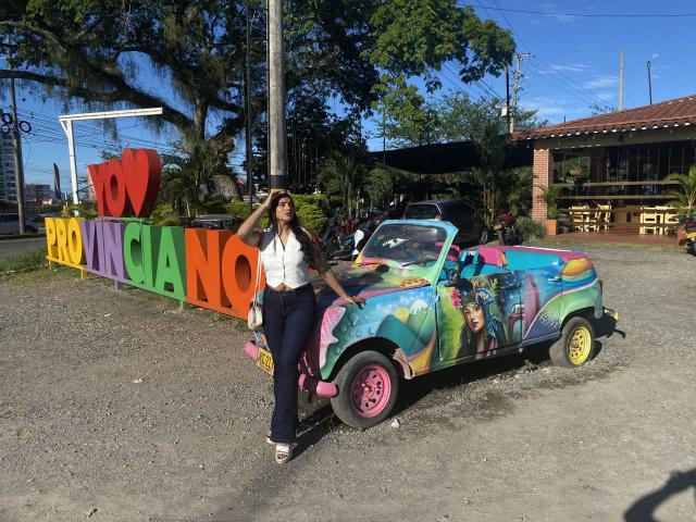 AbbyKell stands in front of a colorful, painted car with "I LOVE CANCUN" in large, multicolored letters. She's wearing a white blouse, navy pants, and black shoes. The car's vivid paint features a mix of abstract and pop art styles, with bright colors and a mix of images, including a person. The car's wheels are pink and yellow. AbbyKell's right hand rests on her head, and she's looking at the camera. The ground is gravelly. The image is brightly lit with clear blue skies.