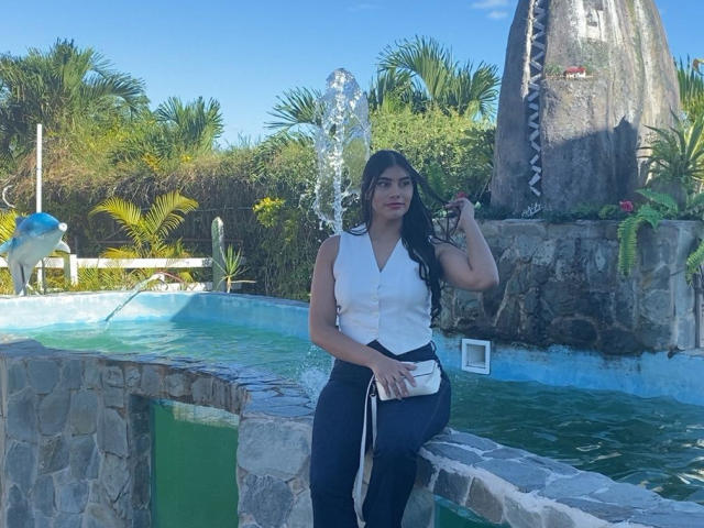 AbbyKell sits by a stone-bordered pool, wearing a white sleeveless button-up shirt and black pants. She holds a small object in her right hand and a white crossbody bag hangs from her left. Her long black hair is loose. A tall rock structure with a cactus and small plants is in the background. A blue and white bird sculpture is to the left. The pool's water is greenish-blue. Sunlight casts shadows.