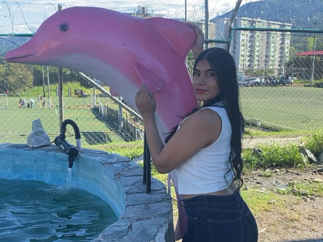 Abby Kell, com cabelos longos e pretos, blusa branca sem mangas e calças jeans escuras, segurando uma estátua rosa de um golfinho, em pé ao lado de uma fonte de água azul, ao ar livre em um dia ensolarado..