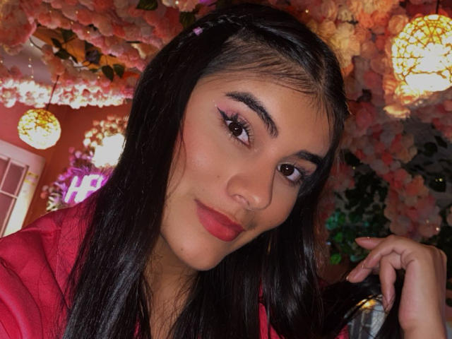 Close-up photo of AbbyKell with medium brown skin, dark straight hair, and red lipstick. She has bold, black eyebrows, thick eyelashes, and pink eyeshadow. AbbyKell wears a red shirt, with her right hand touching her hair. The background has pink floral decorations and warm, yellow lights. Her expression is slightly smug, with a subtle smile. The image has a soft, warm color palette. She looks directly at the camera with a confident gaze.