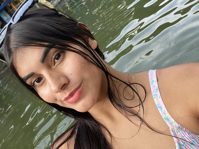 Close-up photo of AbbyKell. She has wet, dark brown hair, light brown skin, and brown eyes. She wears a floral strap dress with a pink and white pattern. Her lips are pink and slightly parted. She has a small gold necklace. Her eyebrows are thick and dark. The water in the background is green and rippled. Her hair is partially covering her right ear. The image has a casual, natural look.