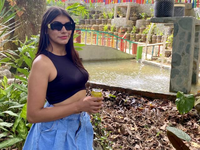 AbbyKell stands in a lush, green outdoor setting, wearing black sunglasses and a black crop top that reveals a small, flat midsection. She holds a yellow drink in her right hand. AbbyKell has long, straight brown hair and light brown skin. She wears high-waisted, light blue jeans. The background features a small stone bridge over a water stream with colorful railings. The image has a casual, relaxed vibe.