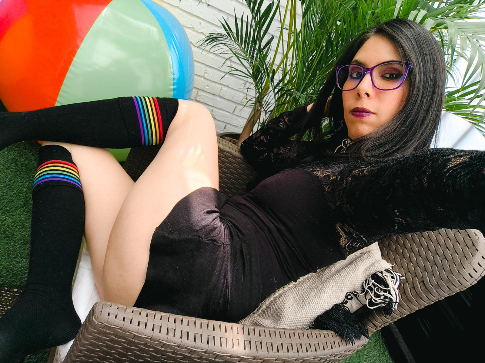 LilithSuccubus with glasses, black lace top, and black shorts, lying in a wicker chair, knees up, rainbow-striped socks, black knee socks, and a colorful inflatable ball in the background.