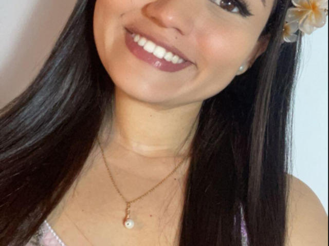 NaughtyxBunny has long dark brown hair, a flower in her hair, a gold necklace with a pearl, and is smiling with pink lipstick and black eyeliner.