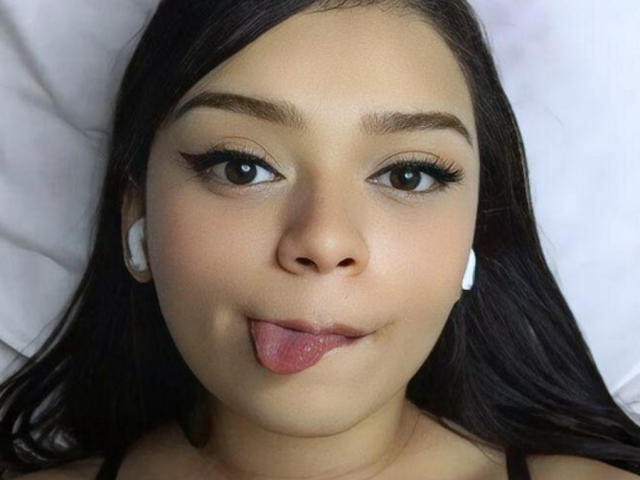 NaughtyxBunny's face, sticking out her tongue with long eyelashes, white earbuds, dark straight hair, and brown eyes, looking cute and playful.