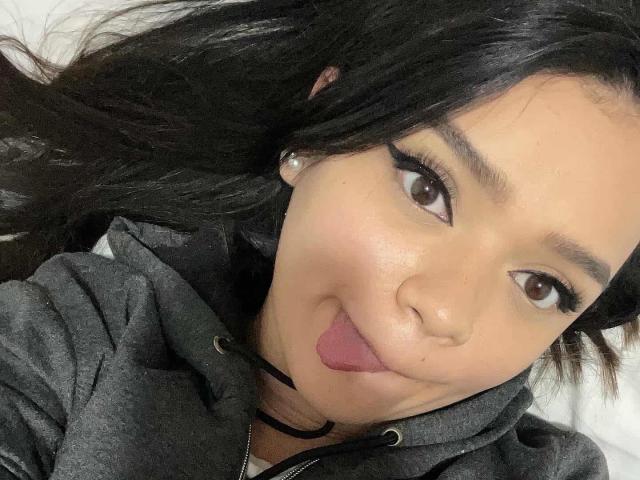 NaughtyxBunny mit langen, schwarzen Haaren, braunen Augen und schwarzem Eyeliner, die ihre Zunge herausstreckt, trägt eine graue Kapuzenjacke, Perlenohrringe und eine schwarze Halskette..
