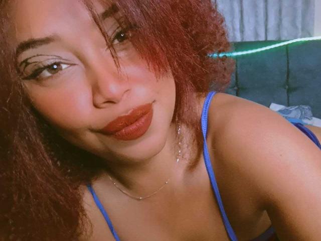 DalilaHeinz with curly red hair, brown skin, and full red lips, wearing a blue spaghetti strap top, smiling softly at the camera, showing her bare shoulders.