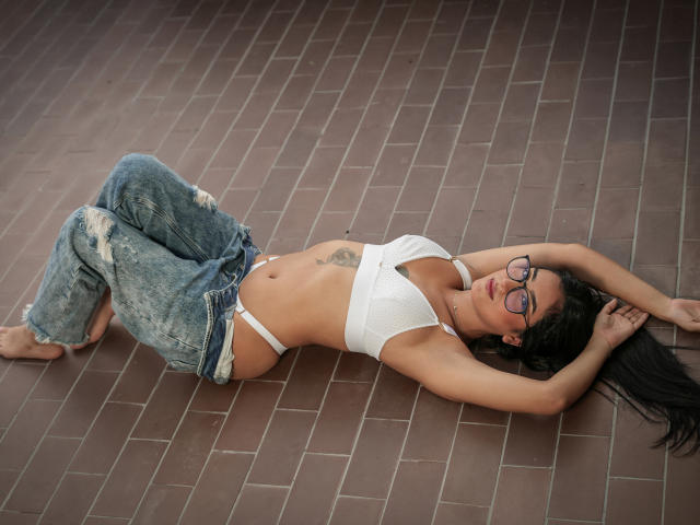 LunaTtorres lies on a brown tiled floor, arms above her head, wearing a white bralette and ripped blue jeans. She has long black hair, glasses, and a small tattoo on her right side. Her pose is relaxed, legs slightly bent. The photograph emphasizes her slim, toned physique and light brown skin. The image has a casual, candid vibe.