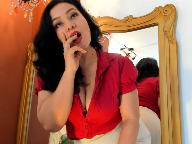 AlisGourmande, avec ses cheveux noirs ondulés, sa chemise rouge à boutons et sa jupe blanche, a un doigt dans la bouche et montre sa poitrine, son reflet se voyant dans un miroir doré orné. Elle a les lèvres rouges et une expression séduisante..