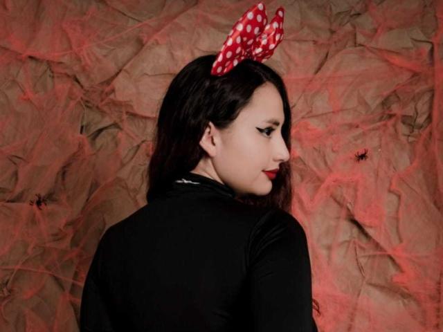 KatiaJamis con un fiocco rosso a pois, una camicia nera a maniche lunghe, rossetto rosso e capelli scuri, che guarda oltre la spalla, su uno sfondo marrone con una luce rossa, e un piccolo ragno nero..