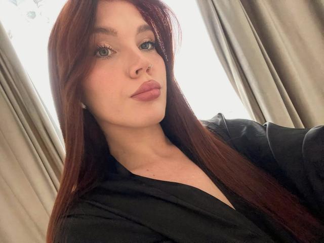 AngelSweetShy, com cabelos vermelhos longos, piercing no nariz e lábios rosados cheios, vestindo uma camisa preta, tem pele clara e olhos verdes, olhando para a câmera com um leve sorriso..