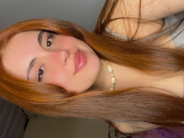 VicluX with long red hair, wearing a gray top, has pink glossy lips, brown eyes, and a shell necklace. Her tits are slightly visible under the top.
