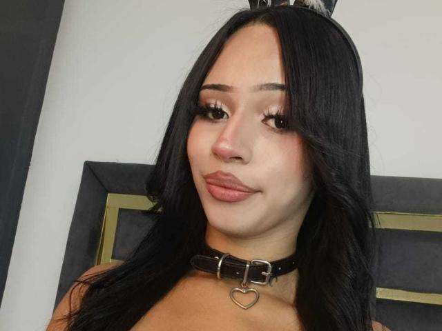 MaxxineVx, con cabello largo y negro, lleva un collar de cuero marrón con una hebilla en forma de corazón, tiene labios carnosos y pestañas postizas, y mira ligeramente hacia la derecha, sonriendo sutilmente..