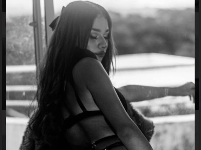 Fotografia in bianco e nero di MaxxineVx con lunghi capelli scuri, che indossa un reggiseno nero, mentre guarda oltre la spalla, mostrando la schiena e i glutei, in piedi vicino a una finestra..
