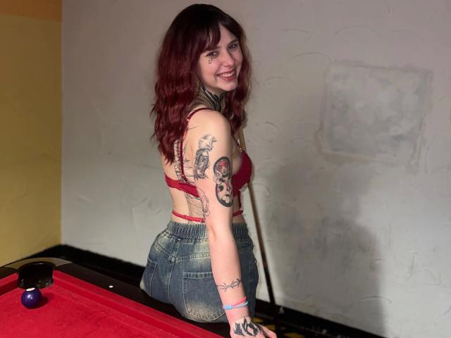 Teufelshelferin-hot with red bra, tattoos, and denim shorts, smiling at camera, standing by red pool table, pool cue behind. Her ass is visible, she has a cute face.