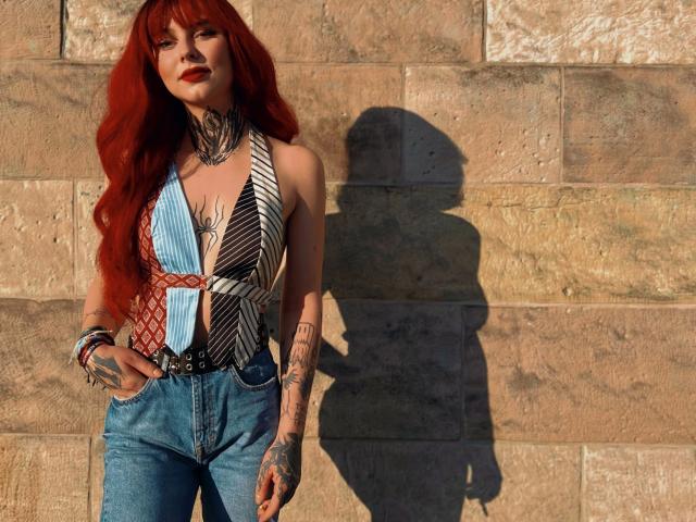 Teufelshelferin-hot with red hair, tattoos, deep cleavage in a patterned top, blue jeans, and red lipstick, standing against a stone wall, shadow on the wall.