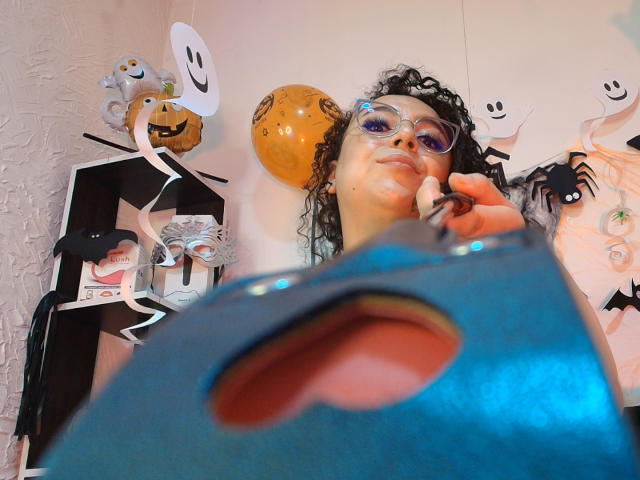 NatashaAmbeer with curly hair, glasses, and a serious expression, wearing a blue mask with a mouth hole, surrounded by Halloween decorations including a smiling jack-o'-lantern balloon.