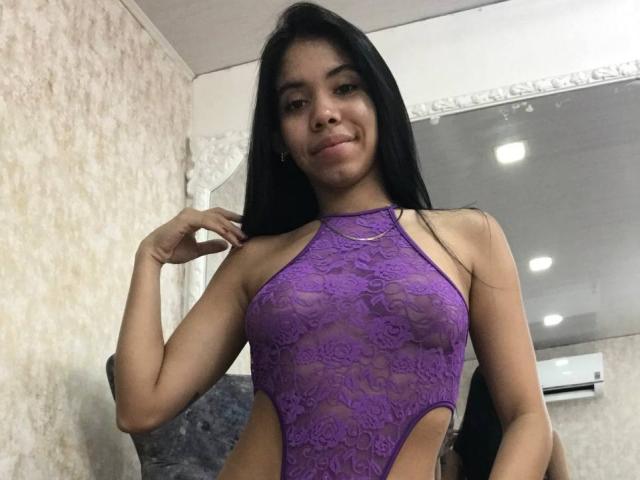 Ema Garces, con lunghi capelli neri, sorridente, indossa un top viola in pizzo con spalline che mette in mostra il suo seno piccolo e presenta dei tagli laterali, ed è in piedi in una stanza con uno specchio..