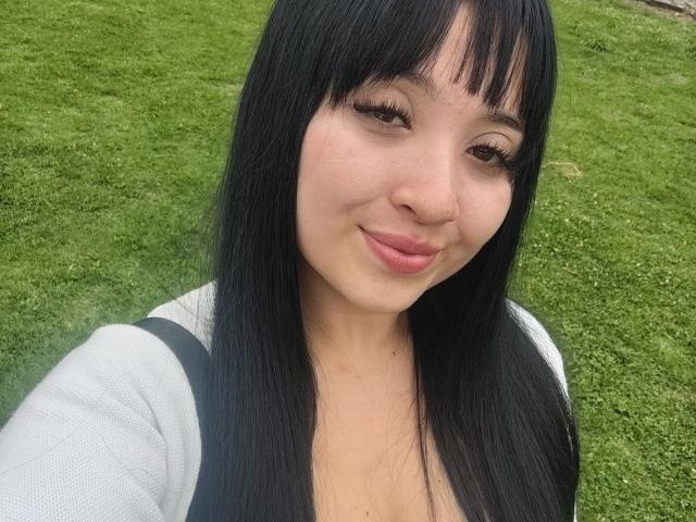 LunaBellaHotty has black straight hair, bangs, and a cute smile with pink lips. She's wearing a gray top, showing a small cleavage. Green grass background.