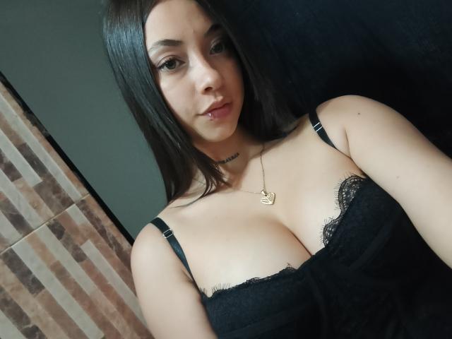 LunaUrbanoo with medium tits in black lace lingerie, black straps, brown hair, subtle makeup, gold necklace, black background, and a slightly parted lip.