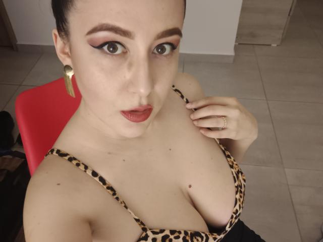 MabyLegrand with dark hair, bold eyeliner, red lipstick, and gold earrings, wearing a leopard-print bra, showing her big, fluffy tits, hand near chest, ring on finger.