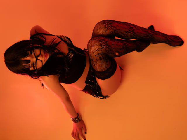 MillaHottLove69 with black lace lingerie, fishnet stockings, and glasses, sitting on her knees, hands on the floor, showing her ass and black lace panties. Orange lighting.
