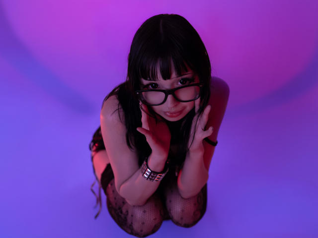 MillaHottLove69 kneels, black-framed glasses, hands on cheeks, smiling, dark hair, pink-purple lighting, black lace stockings, wrist bracelet, looking up, seductive, cute.