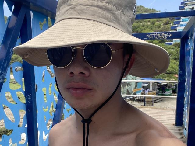 Close-up of SteveWolf. Wearing a beige bucket hat, round sunglasses with a black cord, and no shirt. Sunlight reflects off the sunglasses. SteveWolf's face is centered, with a neutral expression. Background features a blue metal structure with cut-out patterns and a coastal area. The image has a casual, summer vibe. SteveWolf's hat casts a slight shadow over his face. The metal structure's blue paint contrasts with the natural green and blue of the background.