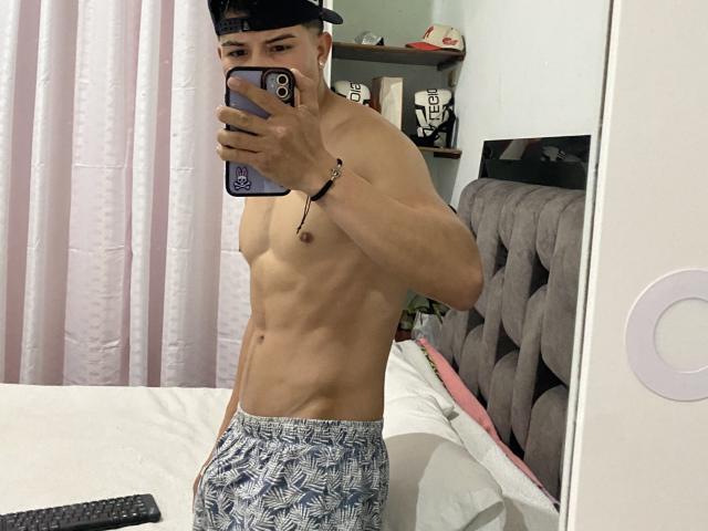 SteveWolf, senza camicia e con un corpo asciutto e tonico, indossa pantaloncini blu con un motivo e un berretto nero, e sta scattando un selfie allo specchio con il suo telefono. Le sue natiche sono leggermente visibili..