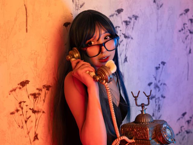 MillaHottLove69 with long black hair, glasses, and a pink top, holding a vintage gold phone to her ear, standing next to an ornate brass phone.
