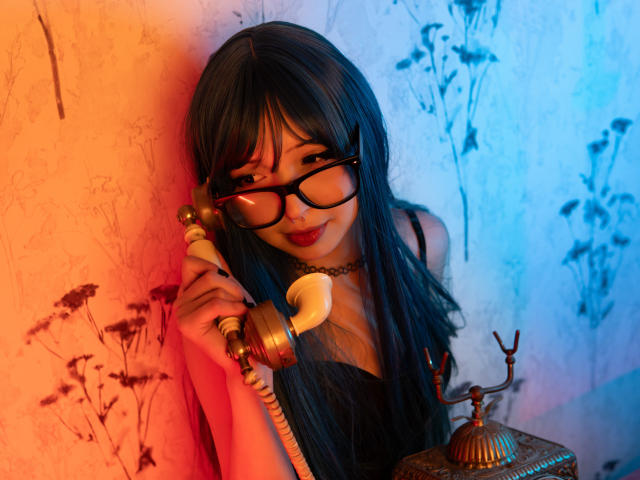 MillaHottLove69, avec de longs cheveux noirs, des lunettes et un joli sourire, tenant un téléphone vintage devant un mur bleu-orange. Sa poitrine est visible sous un haut noir, c'est sexy..