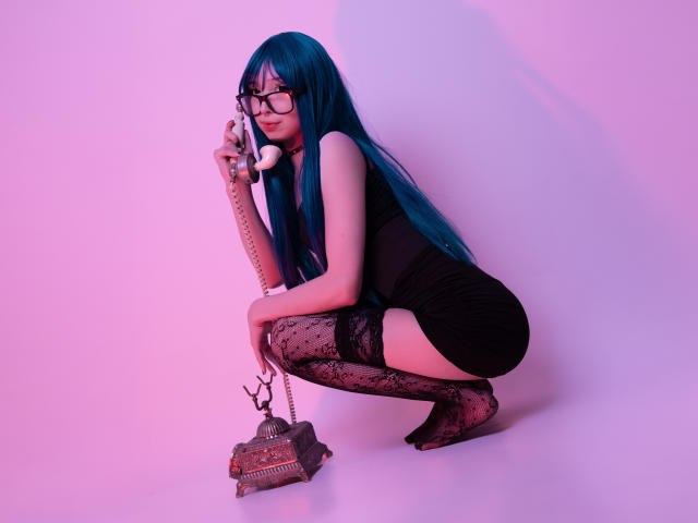 MillaHottLove69 avec des cheveux bleus, des lunettes, une robe noire, des bas en dentelle, en position accroupie, tenant un téléphone, avec un petit sac par terre, éclairage rose et violet. Seins, fesses, vagin..