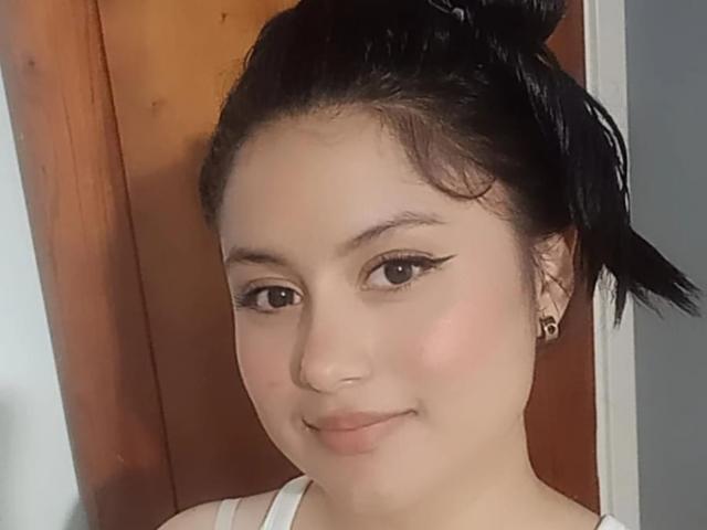 AvaKaine has light brown skin, dark hair in a messy bun, black eyeliner, and a subtle smile. She's wearing a white top with small earrings.