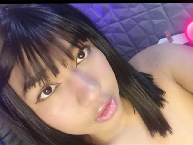 MaddyMagic has glossy black eyes, shiny pink lips, and straight black hair with bangs. Her bare shoulders are visible. She has a cute, sexy face.
