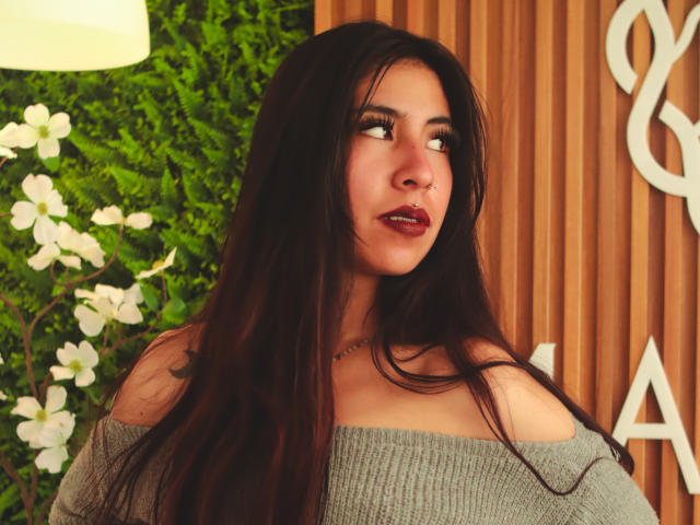 LinaSpearce with long dark brown hair, off-shoulder gray sweater, red lipstick, and nose ring, gazing sideways. Her tits are partially visible under the sweater.