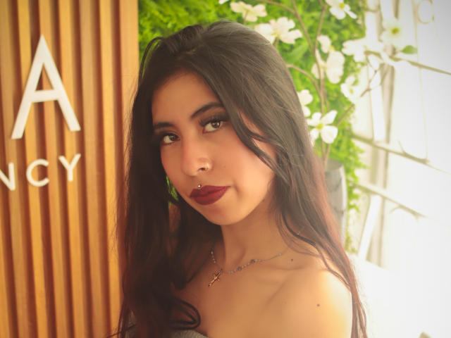 LinaSpearce has dark, wavy hair, brown eyes, and wears red lipstick with a nose ring. She's topless, showing a cross necklace. Her tits are small, and she's smiling.