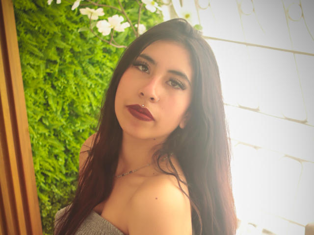 LinaSpearce, avec de longs cheveux bruns foncés, du rouge à lèvres foncé, et un anneau de nez, portant un haut gris à une épaule, regardant la caméra avec un léger sourire..