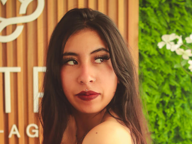 LinaSpearce has dark brown hair, red lipstick, and a nose piercing, looking to the side with half-closed eyes, showing her bare shoulders. Her makeup is bold and sexy.