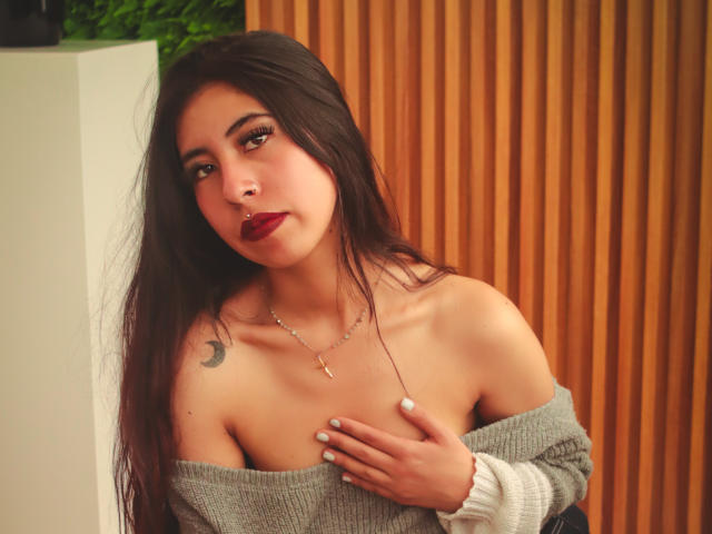LinaSpearce with long dark hair, red lipstick, off-shoulder grey sweater, hand on chest, visible cleavage, small crescent moon tattoo on left shoulder, wooden background.