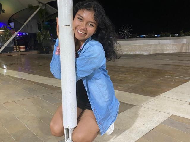JuliaBennett, kneeling, smiling, holding a pole, wearing a blue denim jacket, black shorts, and white sneakers, with curly black hair, and brown eyes, at night.