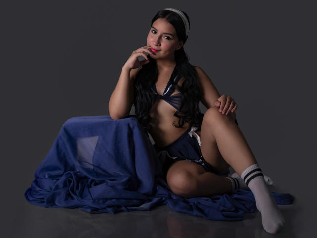 Valentine Rouse, com longos cabelos pretos, sentada em um lençol azul, vestindo um sutiã e shorts azul marinho, meias brancas e uma faixa de cabelo, mordendo o dedo, mostrando os seios..