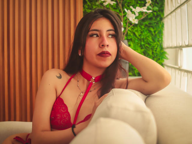LinaSpearce with dark hair, red lace bra, and choker, resting on a white couch, looking to the side, showing a small crescent tattoo on her right shoulder.