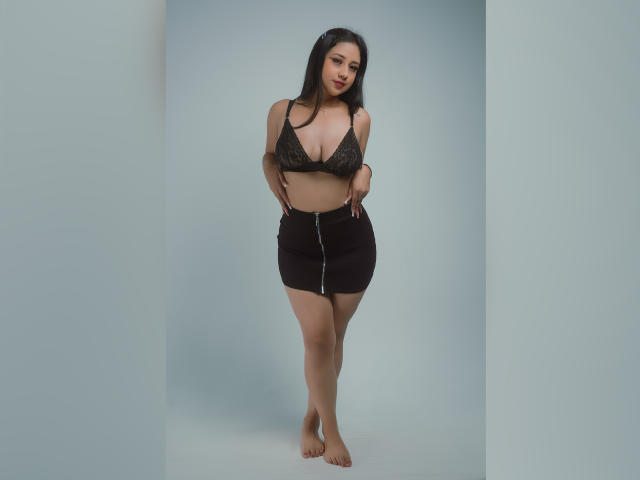ElliePayton with dark hair, wearing a black lace bra, showing her boobs, and a black velvet skirt with a zipper, standing barefoot, hands by her sides, looking down.