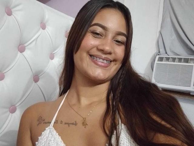 RoxyWilliamss, com cabelos castanhos longos, sorrindo, vestindo uma blusa branca de alças, mostrando uma tatuagem no ombro direito, e um colar com uma pequena cruz dourada..