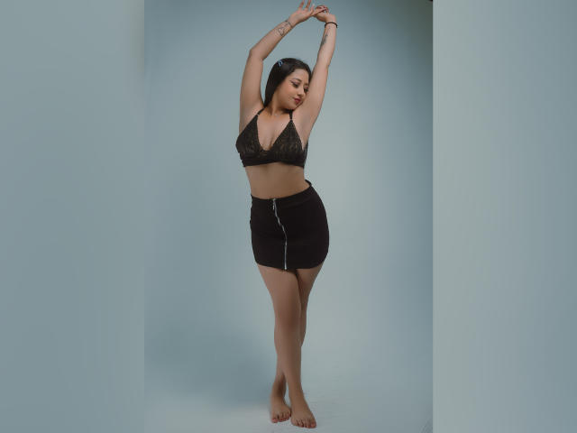 ElliePayton with arms raised, wearing a black lace bra and black skirt with a zipper, standing barefoot, showing her tits and ass. She has tattoos on her arms.