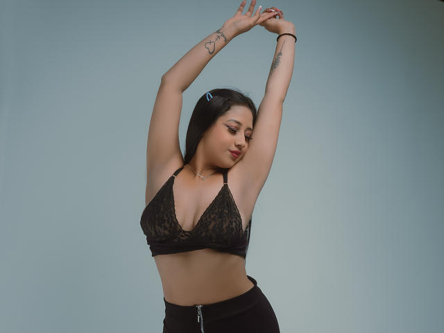 Ellie Payton, con le braccia alzate, indossa un reggiseno in pizzo nero, mostrando il seno, pantaloni neri a vita alta, tatuaggi sulle braccia e una molletta per capelli nei suoi capelli scuri..