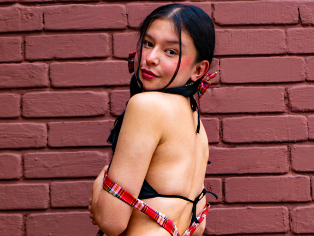 DyanaMoon mit dunklem Haar, trägt einen schwarzen Bikini-Oberteil, rote Karomuster-Armbänder, blickt über ihre Schulter und zeigt ihren nackten Rücken und ihre kleinen, prallen Brüste. Eine rote Backsteinmauer..