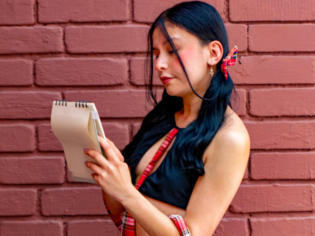 DyanaMoon with black hair, red ribbon, and plaid outfit, holding a notebook, showing her tits through a deep black top, against a red brick wall, looking down.