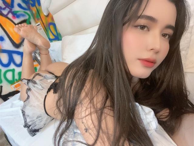 LianaBernocchi with long black hair, lying on a bed in black and white lingerie, her bare ass and feet up, looking at the camera with red lips.